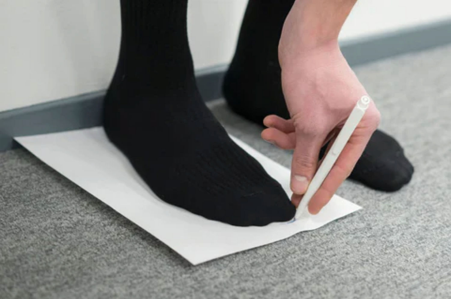 A foot placed on a piece of paper with the heel touching a wall behind. Tip of te foot is marked on the paper with a pen