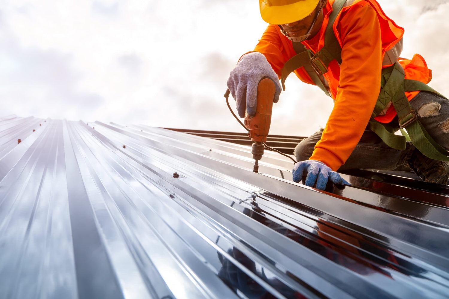 On a metal roof, a construction roofer is wearing his Personnel Protective Equipement (PPE). He could be wearing magnetic boots. He is fixing a screw on a steel panel