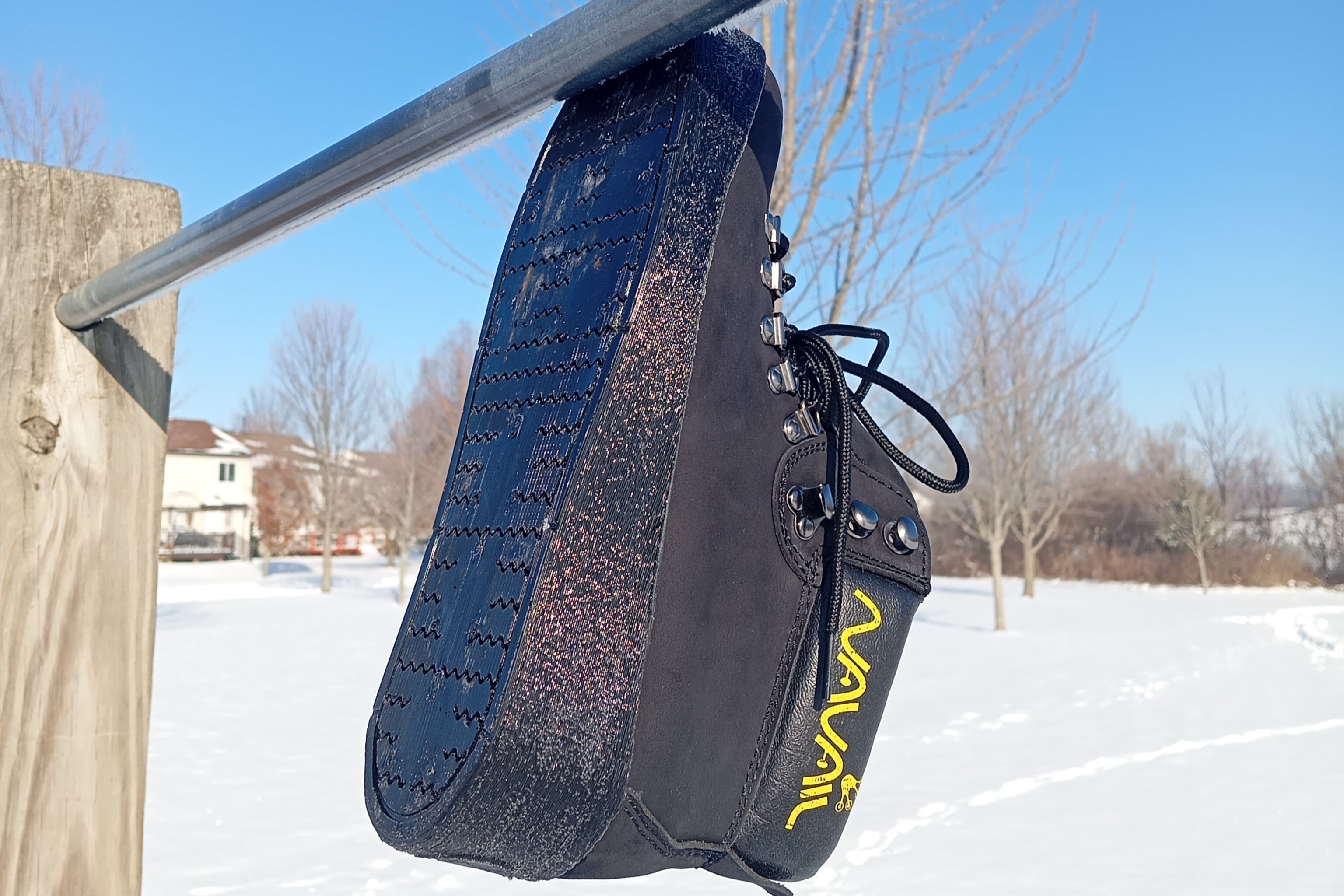 A Magnetic work boot is clung unto a horizontal metal pole. Magnets in the sole make the boot stick to the bar. Snow is visible in the background, suggesting grip even in cold and wet conditions